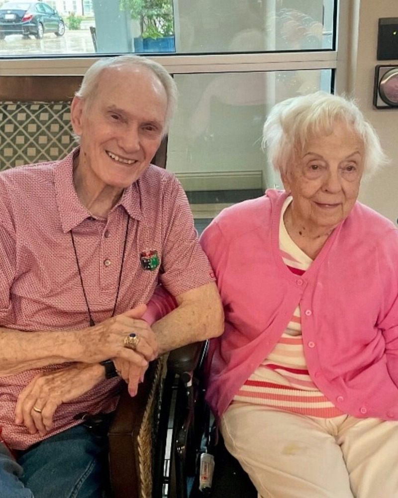 An elderly man and woman sit side by side, smiling warmly together near a bright window.