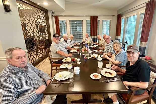 A group of older adults seated together around a long dining table, sharing a meal in a warmly decorated dining room with large windows and elegant room dividers.