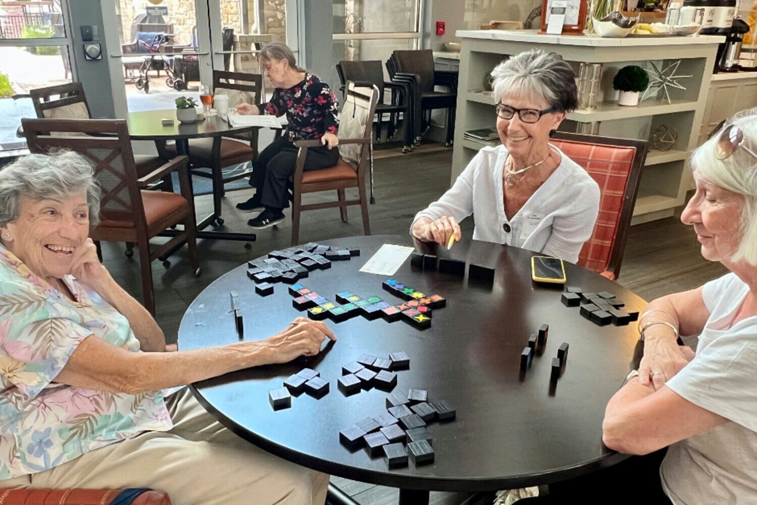 Three older women smile and laugh while playing a dominoes game at a round table in a bright, sunlit common area, with another resident seated in the background.