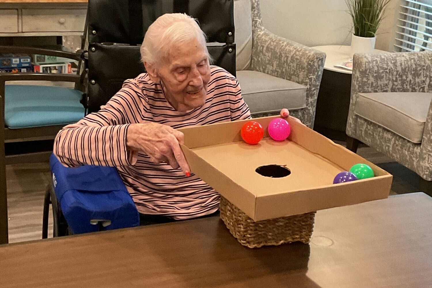 An elderly woman seated at a table playing a tabletop ball-drop activity game with colorful balls and a cardboard box with a hole.