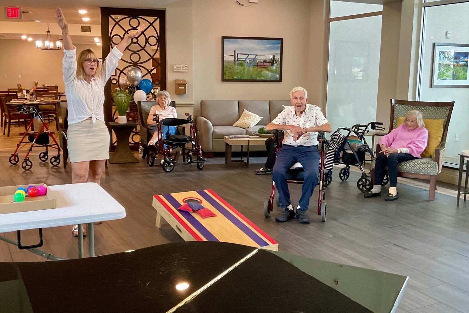 A staff member celebrates while playing cornhole with senior residents in a bright, spacious common room at a senior living community.
