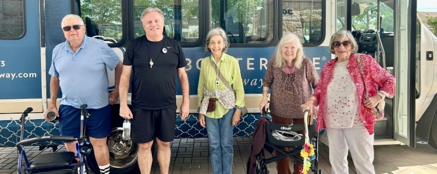 A group of seniors and a staff member smile together in front of a community transportation bus, ready for an outing.