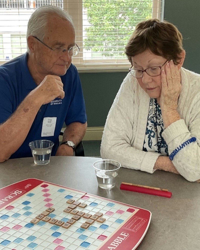 Two seniors sit together at a table, deep in concentration while playing a game of Scrabble.