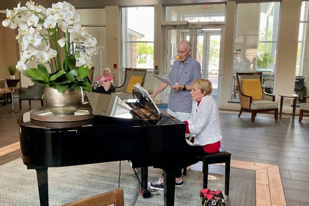 A woman plays a grand piano in an elegant senior living community lobby while a man stands beside her reading sheet music.