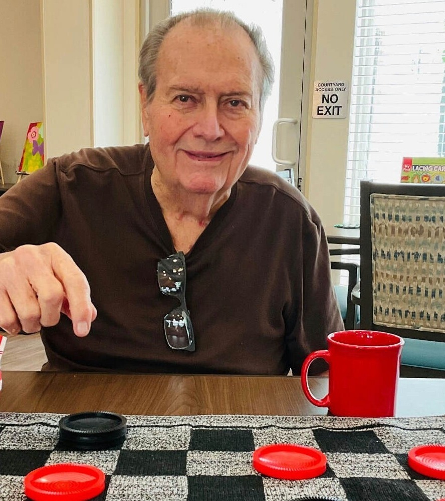 An older man smiles while reaching to make a move in a game of checkers, with a red coffee mug beside the board on a wooden table.