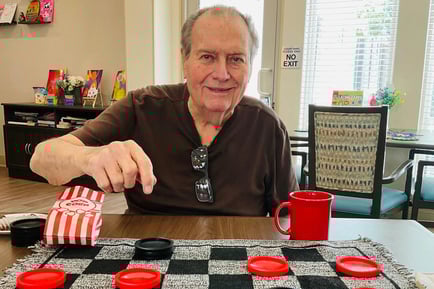An elderly man smiles while reaching across a checkerboard game table, with a red mug and striped cloth nearby. 