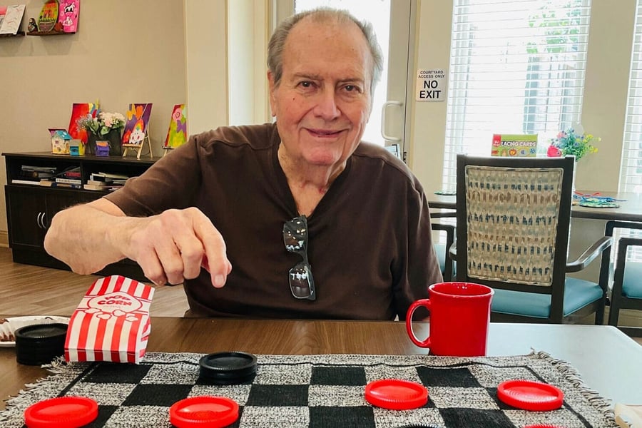 An elderly man smiles while reaching across a checkerboard game table, with a red mug and striped cloth nearby. 