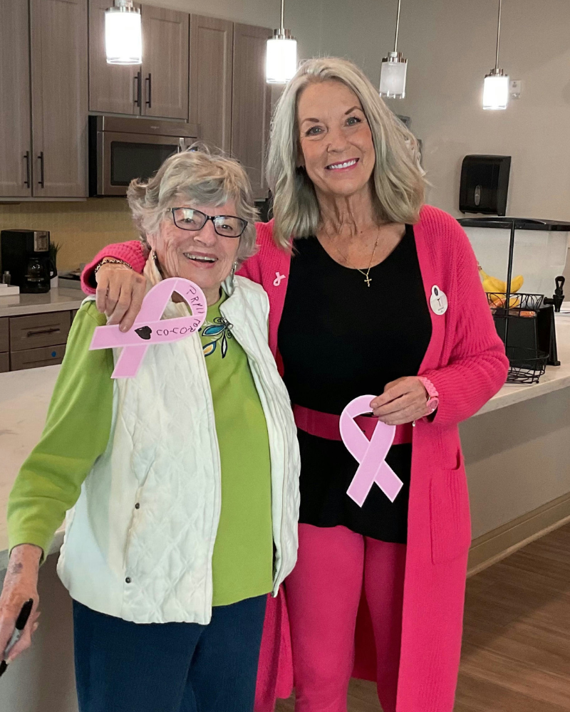 Two women smiling and posing together indoors, each holding a pink ribbon, symbolizing breast cancer awareness.