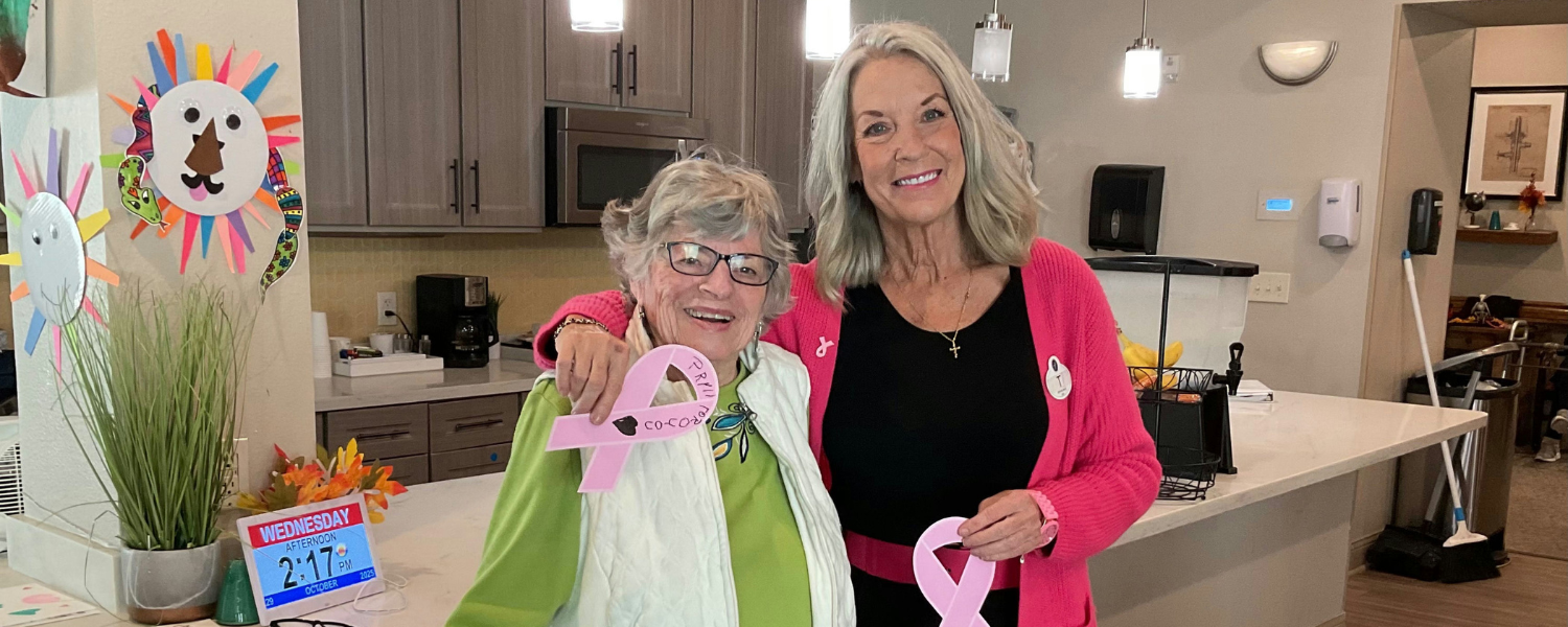 Two women smile and pose together in a kitchen or common area, each holding a pink ribbon in support of breast cancer awareness.