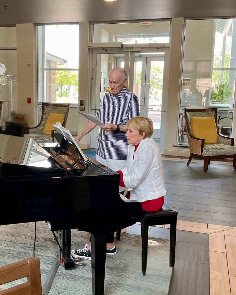 An older man stands beside a grand piano, pointing at sheet music, while an older woman sits and plays in a bright, airy common room.