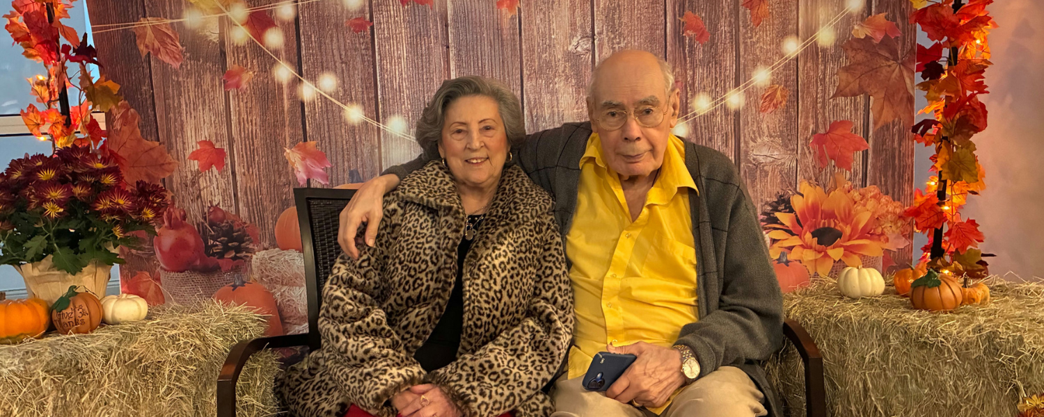 Two seniors pose together in front of a festive fall-themed backdrop at Arbor Terrace Lakeway, surrounded by autumn leaves, pumpkins, hay bales, and warm string lights at this senior living community in Lakeway, TX.