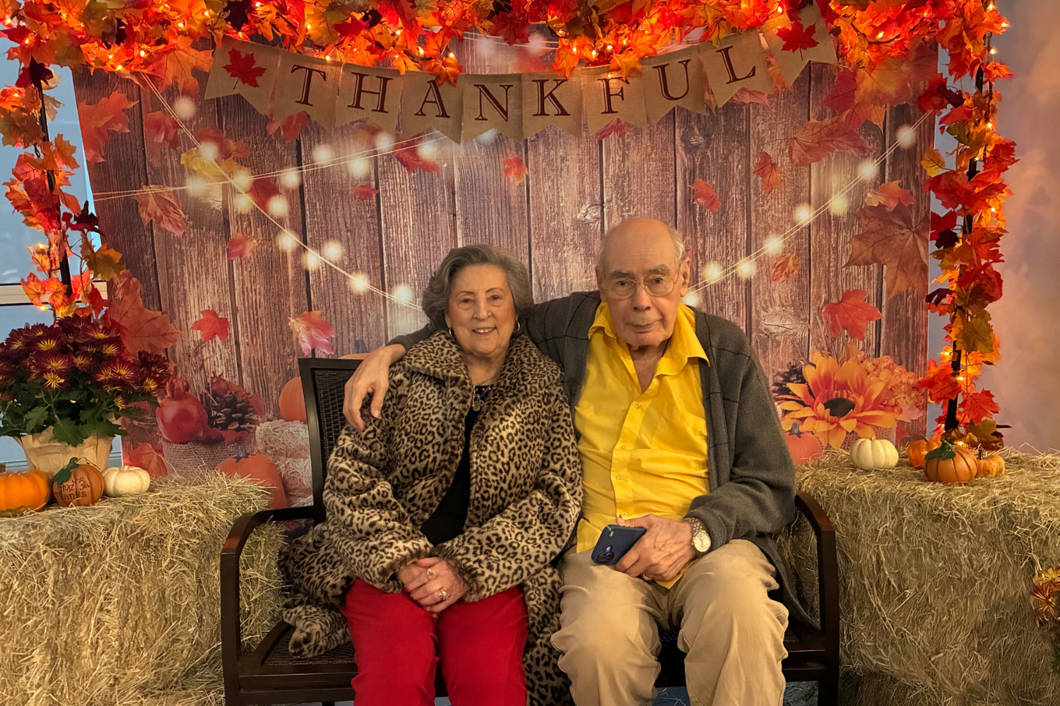 Two residents sit together in front of a festive "Thankful" Thanksgiving backdrop with autumn décor and pumpkins at Arbor Terrace Lakeway TX, celebrating the holiday season at this senior living community.