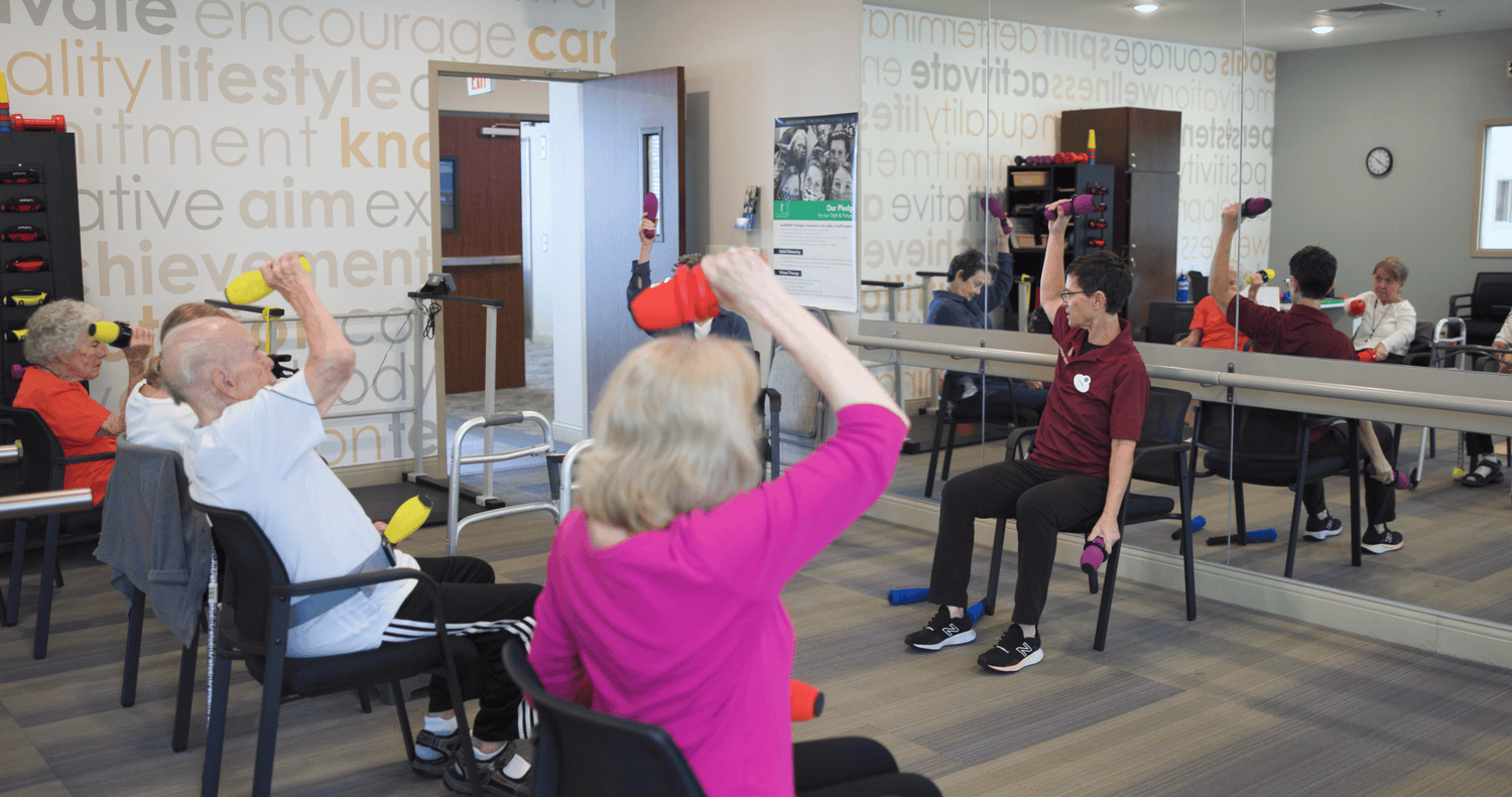 A group of seniors participates in a seated exercise class, lifting hand weights with an instructor in a fitness room decorated with motivational words on the walls.