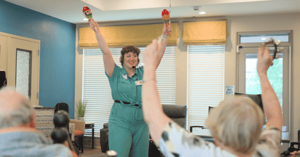 A staff member leads an energetic group activity at Arbor Terrace Lakeway, holding colorful maracas with arms raised as seniors join in — supporting brain health programs for seniors in Lakeway, TX.