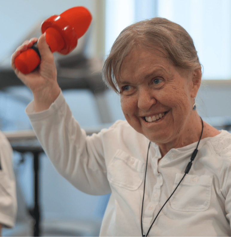 An older woman smiles broadly while lifting a red hand weight during a fitness class, with exercise equipment visible in the background.