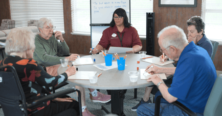 A staff member leads a small group of seniors in a structured table activity in a cozy common room at Arbor Terrace Lakeway — meaningful connection through senior living Lakeway Texas.