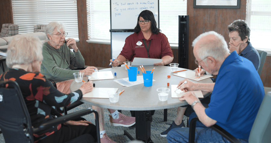 A staff member leads a small group of seniors in a structured table activity in a cozy common room at Arbor Terrace Lakeway — meaningful connection through senior living Lakeway Texas.