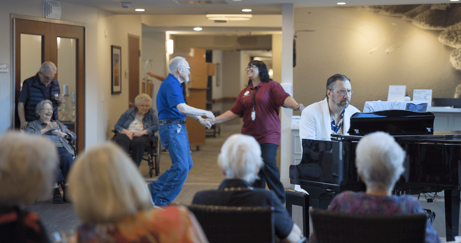 A group of seniors gather in a common area to enjoy a live music event, with a man playing a grand piano, a staff member dancing with an elderly resident, and several residents seated watching the performance.