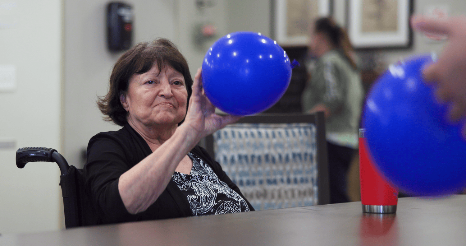 A senior woman in a wheelchair participates in a group balloon activity, reaching out to catch or tap a blue balloon during a recreational activity session.
