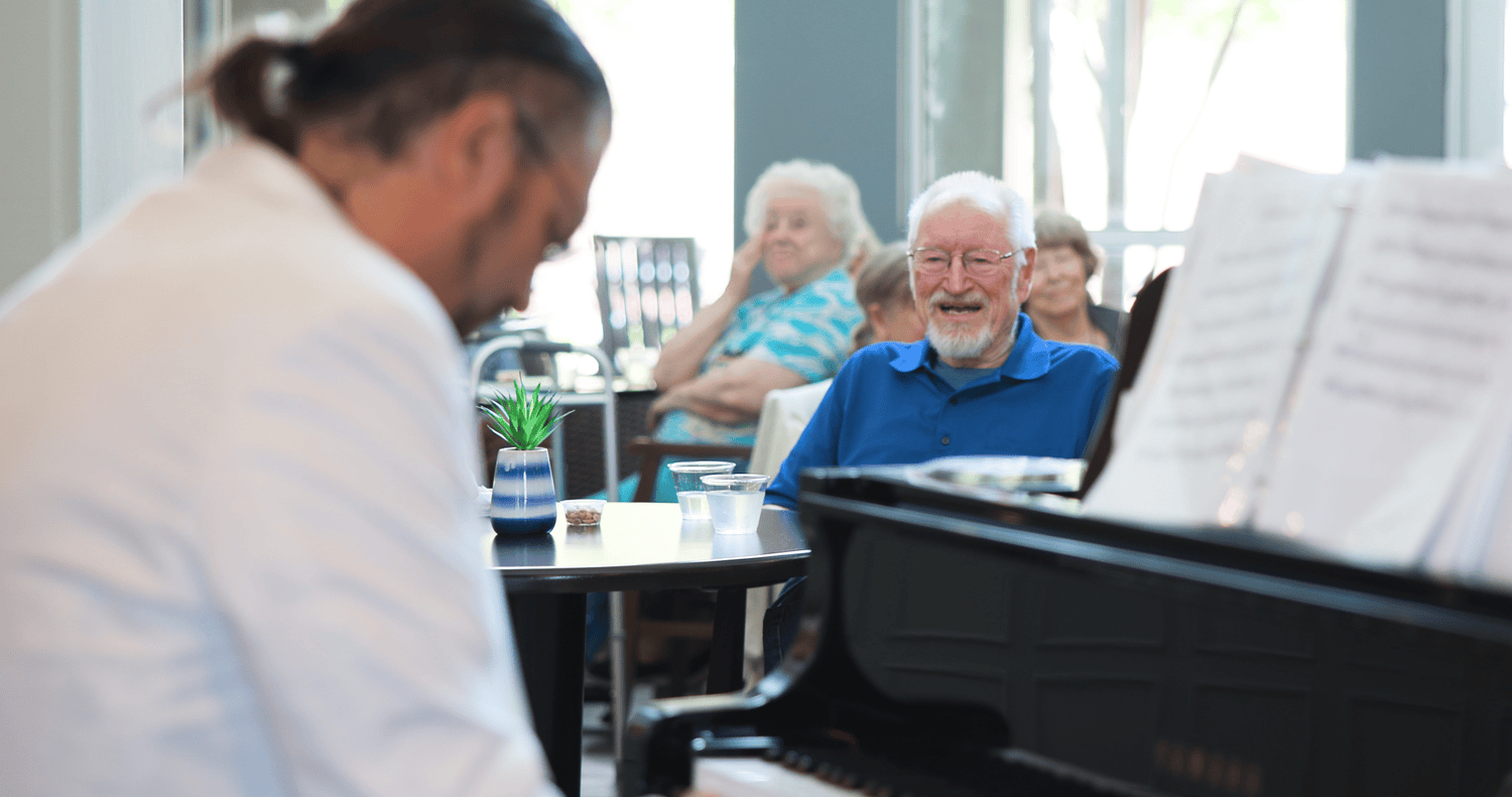 A pianist performs for a group of smiling seniors seated in a bright, elegant common area, with one man in the foreground beaming with enjoyment.