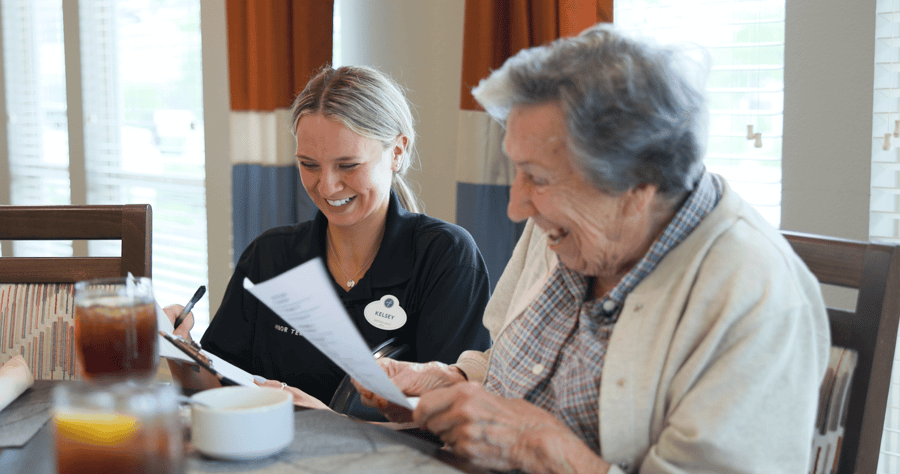 A smiling staff member sits with an older adult at a dining table, reviewing paperwork together at Arbor Terrace Lakeway — warm, personalized senior care in Lakeway, TX.