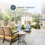 Sunny outdoor patio at Arbor Terrace Lakeway featuring comfortable lounge chairs with blue and yellow cushions around a small table, surrounded by lush trees and a stone building exterior.