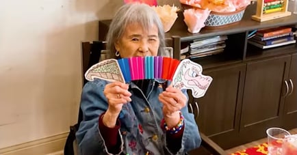 A cheerful older adult holds up colorful craft cutouts and markers at Arbor Terrace Lakeway, beaming with joy — celebrating joyful senior living Lakeway Texas.