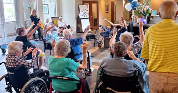 A group of elderly residents at a senior living facility participate in a seated exercise or movement class, raising their arms while an instructor leads them. Several participants are in wheelchairs, and blue and white balloons decorate the bright, cheerful room.