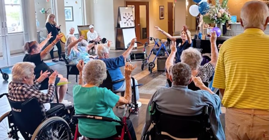 A group of elderly residents at a senior living facility participate in a seated exercise or movement class, raising their arms while an instructor leads them. Several participants are in wheelchairs, and blue and white balloons decorate the bright, cheerful room.
