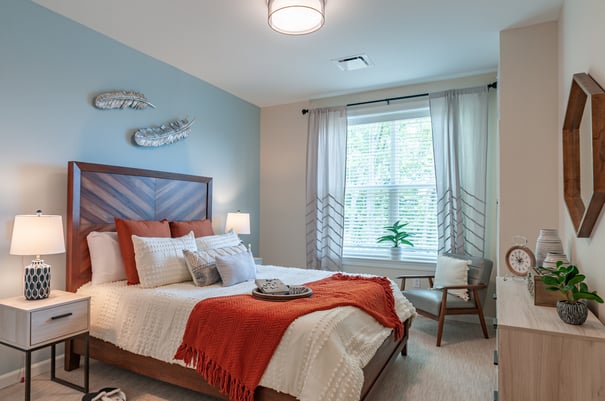 A cheerful, thoughtfully decorated bedroom at this assisted living community in Marlton NJ, featuring a warm wood headboard, a bright orange throw, layered white pillows, a soft blue accent wall with feather wall decor, and a sunny window with sheer curtains.