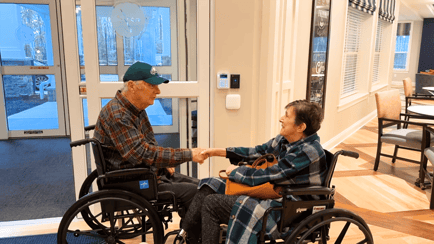 Two elderly residents in wheelchairs shaking hands and smiling at each other inside a bright, modern memory care community.