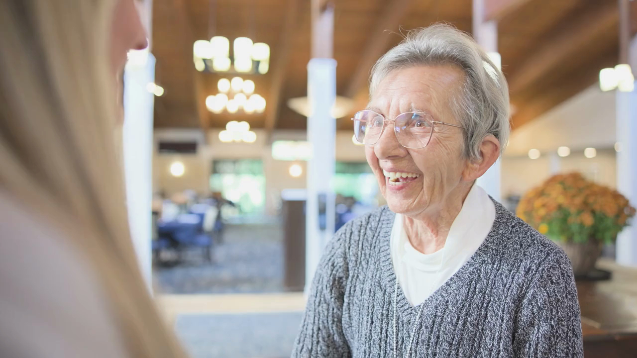 A smiling older woman with glasses chats warmly with another person inside a bright, elegantly appointed common area at this senior living Middletown NJ community, with soft lighting and floral accents in the background
