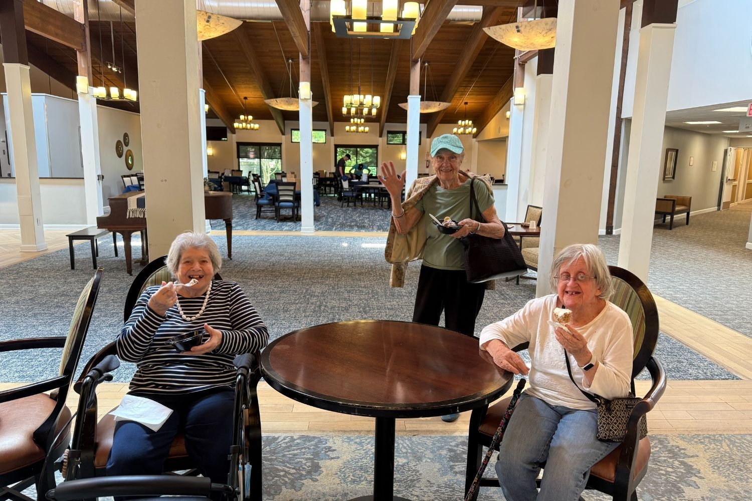 three residents/women smile and gather around a table to eat ice cream. one woman is standing and the other two are sitting