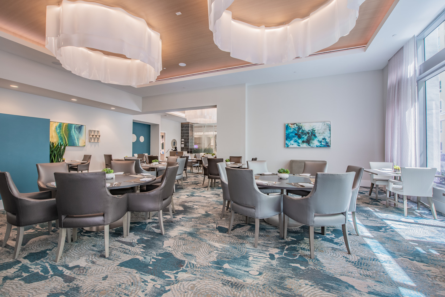 A beautifully appointed dining lounge at Mirabelle in Miami, FL, with round tables, cushioned chairs, a statement chandelier, and a vibrant patterned rug for resort-style senior living.