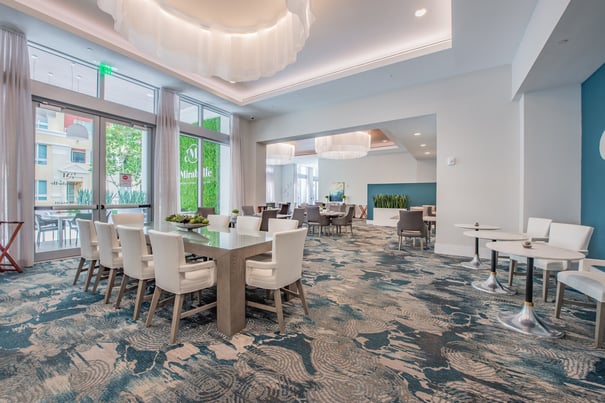 The elegant dining room at Mirabelle features sculptural ceiling details, white upholstered chairs, and large windows, offering a sophisticated on-site dining experience for senior living in Miami, FL.