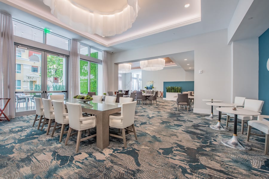 The elegant dining room at Mirabelle features sculptural ceiling details, white upholstered chairs, and large windows, offering a sophisticated on-site dining experience for senior living in Miami, FL.