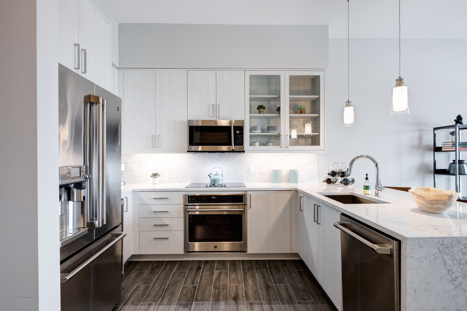 A bright, modern kitchen in a Mirabelle senior apartment in Miami, FL, with white cabinetry, glass-front upper cabinets, stainless steel appliances, and pendant lighting for independent living.