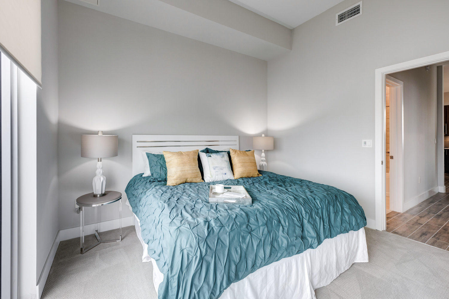 A cozy bedroom in a Mirabelle senior apartment near Pinecrest, FL, featuring a teal bedspread, accent pillows, a wall-mounted TV, and soft ambient lighting for comfortable senior living.