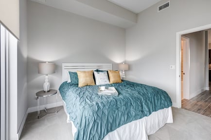 A cozy bedroom in a Mirabelle senior apartment near Pinecrest, FL, featuring a teal bedspread, accent pillows, a wall-mounted TV, and soft ambient lighting for comfortable senior living.