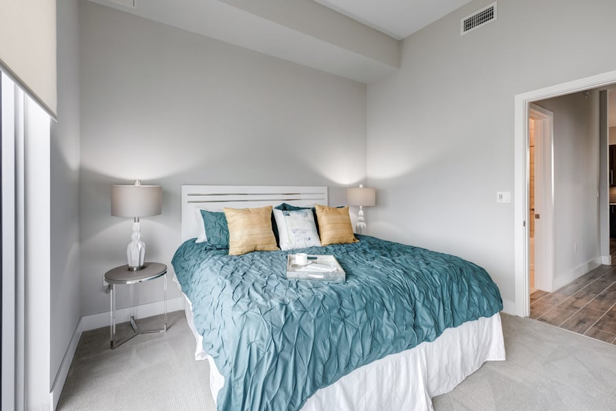 A cozy bedroom in a Mirabelle senior apartment near Pinecrest, FL, featuring a teal bedspread, accent pillows, a wall-mounted TV, and soft ambient lighting for comfortable senior living.