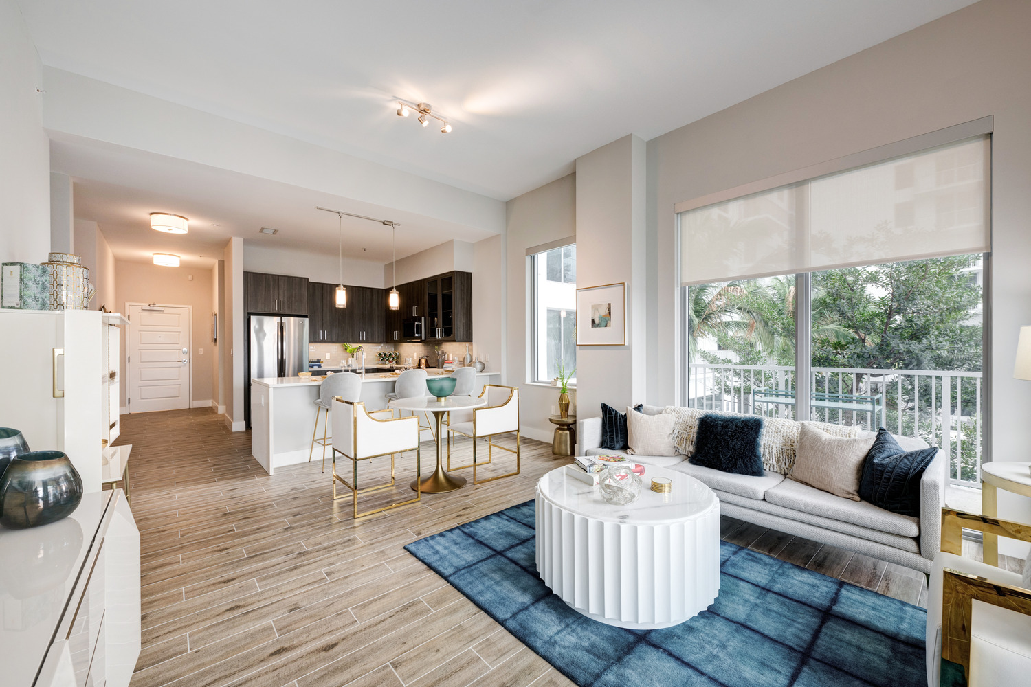 An open living and dining area in a Mirabelle senior apartment near Pinecrest, FL, with a round coffee table, blue rug, and large windows overlooking greenery for active senior living.