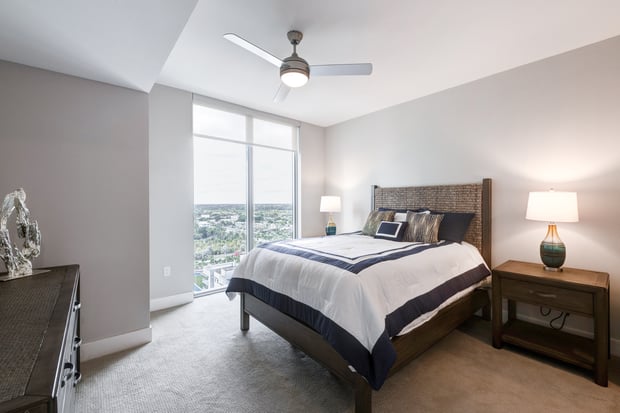 A calm, well-lit bedroom in a Mirabelle senior apartment in Miami, FL, with a wood-frame bed, bedside lamp, ceiling fan, and large windows with sweeping city views for restful living.