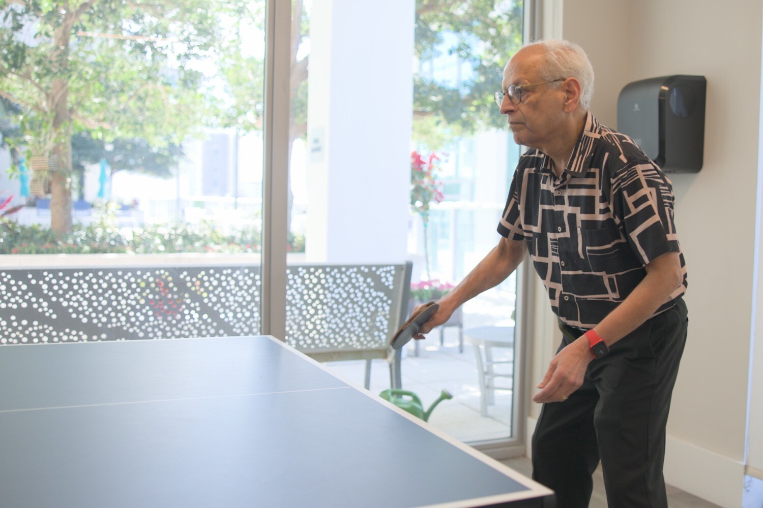 Un residente juega ping pong junto a una ventana con vista a un patio verde, reflejo de la vida sénior activa en Miami, FL.