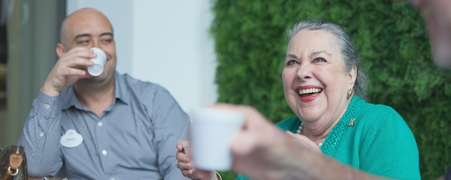 Un miembro del personal y una residente sonríen mientras disfrutan de café al aire libre junto a un jardín verde, parte de la vida sénior activa y la vida sénior en Miami, FL.