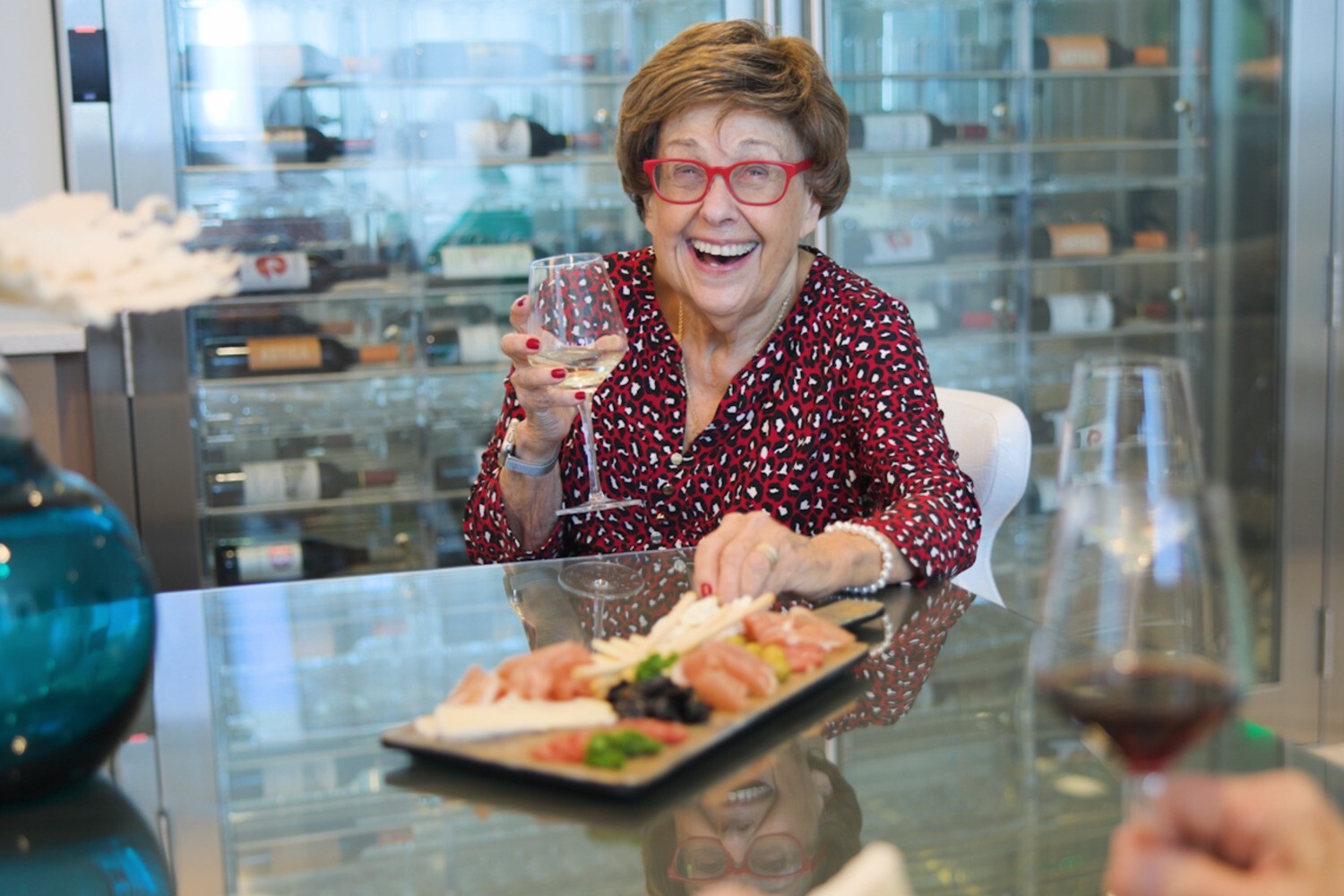 Una residente sonríe con alegría mientras sostiene una copa de vino junto a una tabla de embutidos y quesos, reflejo de la vida sénior con comedor en el lugar y estilo resort en Miami, FL.