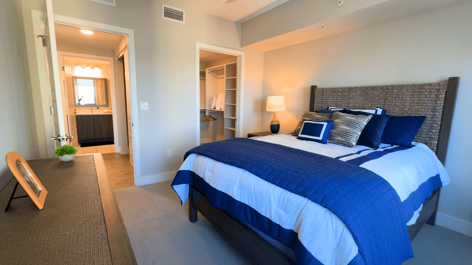 A bright bedroom in a Mirabelle senior apartment in Miami, FL, featuring a navy accent bedspread, a wall-mounted TV, warm wood furniture, and soft natural light for restful independent living.