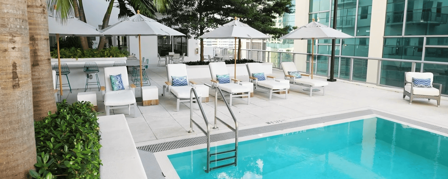 A sparkling outdoor pool with lounge chairs, white umbrellas, and lush greenery at this resort-style senior living community in Miami, FL, perfect for active seniors enjoying the South Miami sunshine.