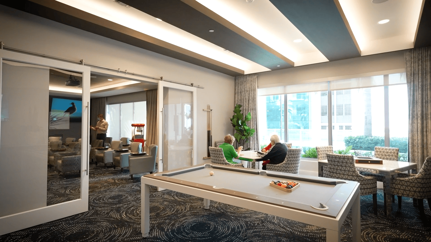 A spacious indoor common area at Mirabelle senior living in Miami, FL, featuring a pool table, modern furnishings, and large windows overlooking the outdoor pool for active senior living.