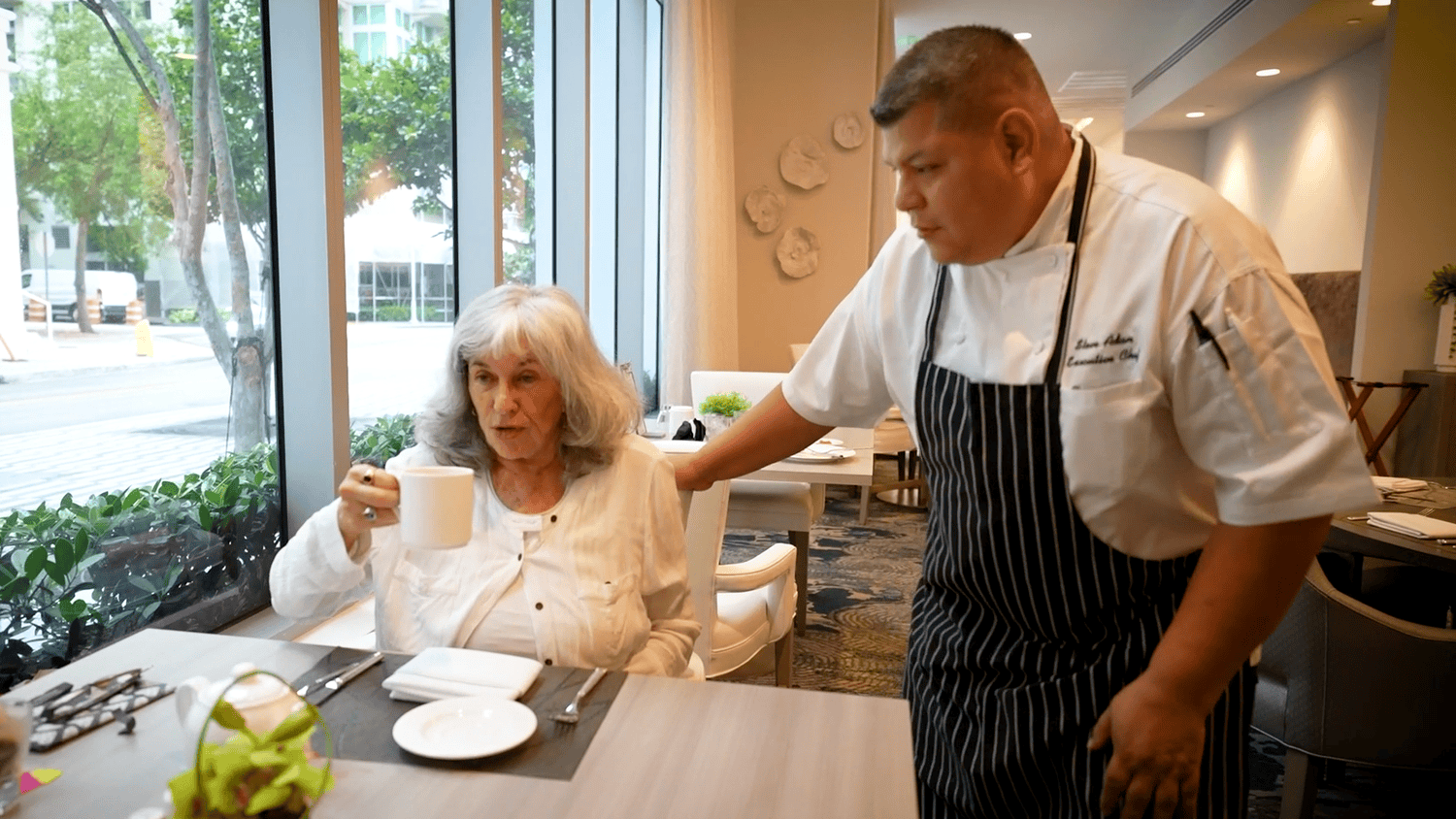 A culinary staff member in a chef's apron serves a resident at Mirabelle senior living in Miami, FL, showcasing the community's commitment to personalized dining experiences.