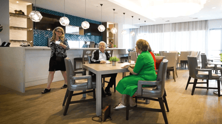 Two residents enjoy conversation at a dining table in a bright, modern common area, while a staff member assists nearby. A welcoming space for independent living in Miami, FL, with pendant lighting and warm wood floors.